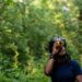 Devin Cowens holds a monocular to her eye, in front of a forested backdrop.