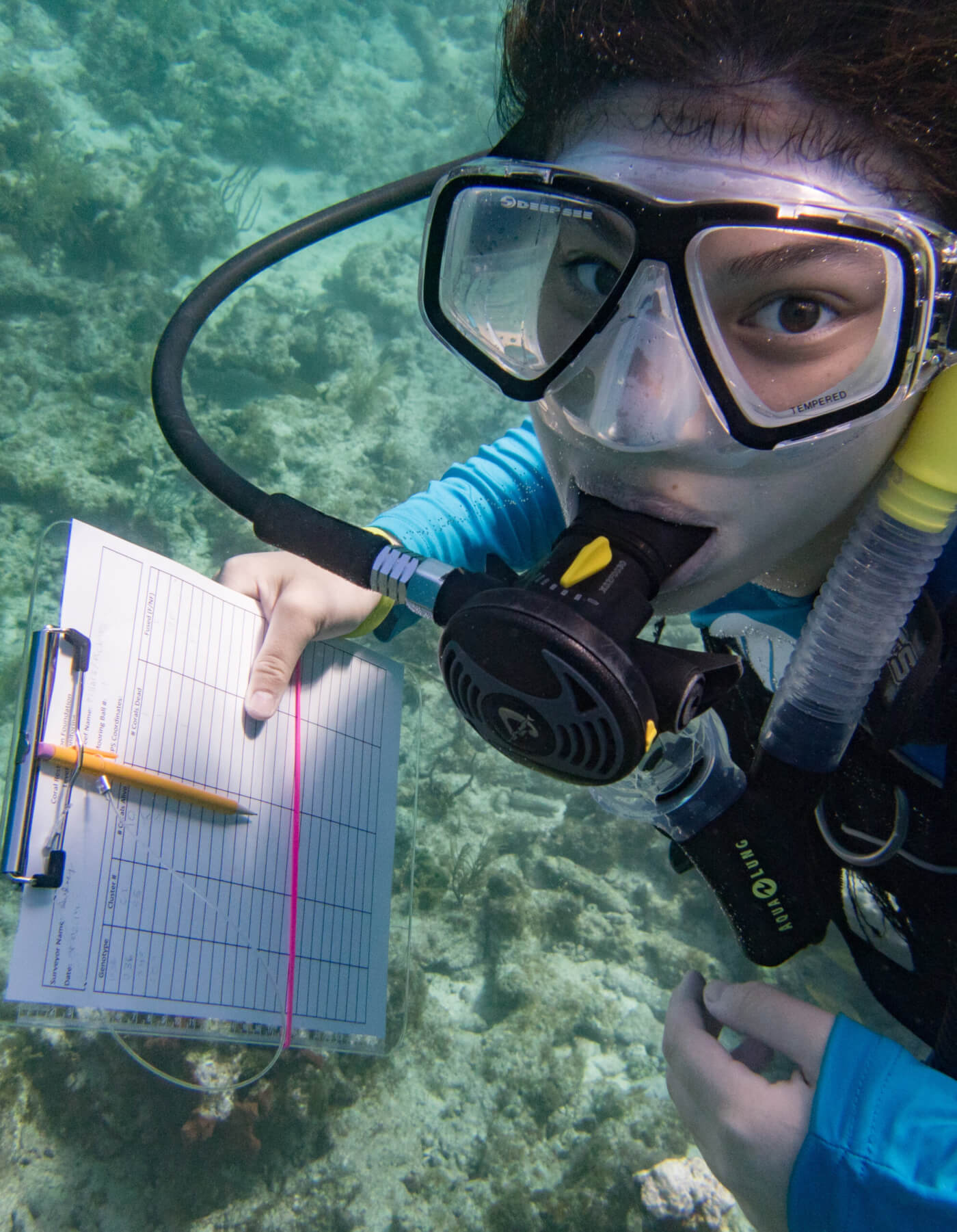 person underwater with scuba gear on looking at camera with clipboard and log chart in hand with reef in background