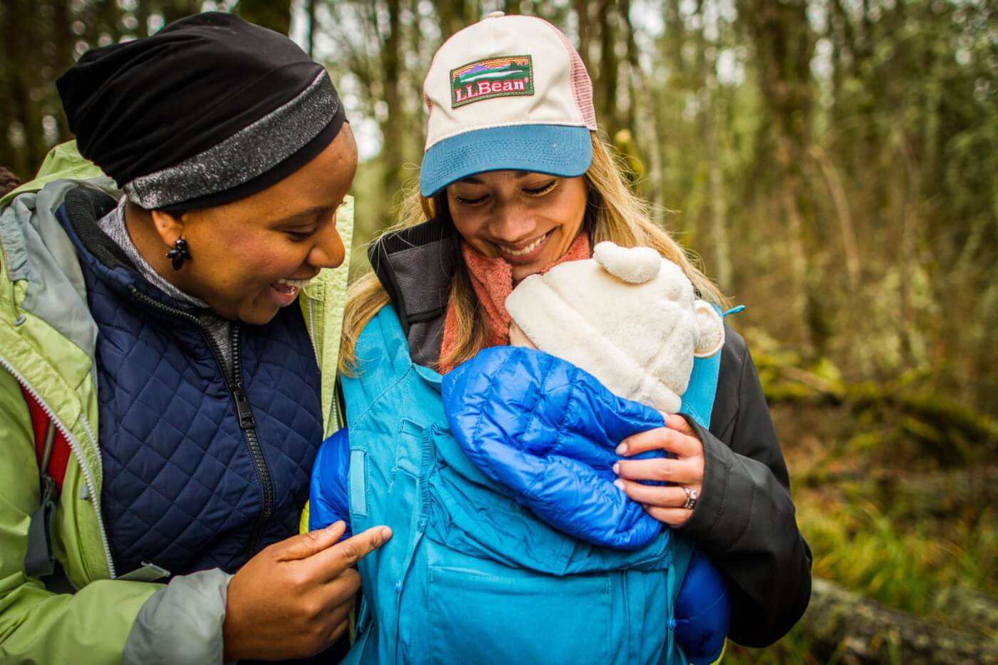two parents going for a hike in the woods with a child in a baby sling on one of their chests. they are both looking down smiling at the child