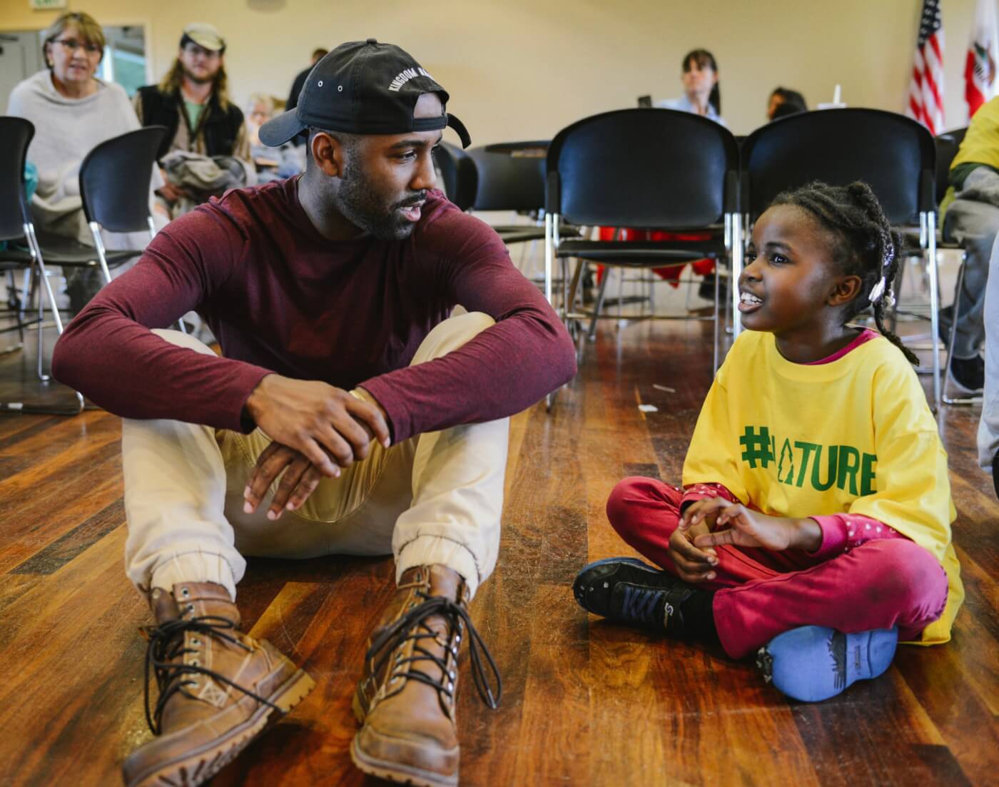 CJ Goulding sits on a wooden floor, in discussion with a young child sitting cross-legged beside him.