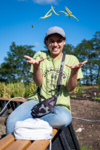 A participant, sitting on a bench with a garden in the background, throws a handful of green beans into the air while smiling.