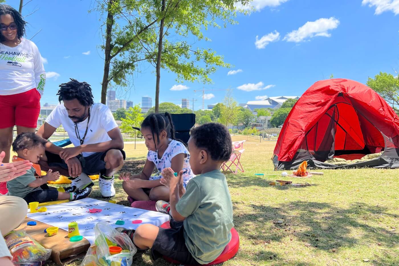 Tent in the middle of a park with children and parents in front of it playing with playdoh. The city is in the background