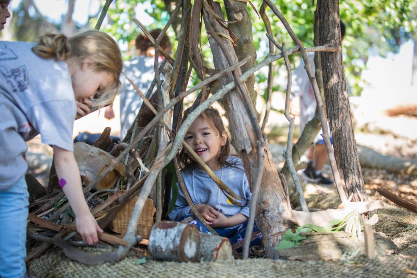 A young child peers through branches of a makeshift wooden structure.