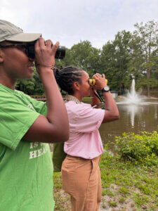 Devin and a friend peer through binoculars in front of a pond and water fountain.