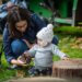 An adult holds a pinecone for a baby wearing a white cap to explore.