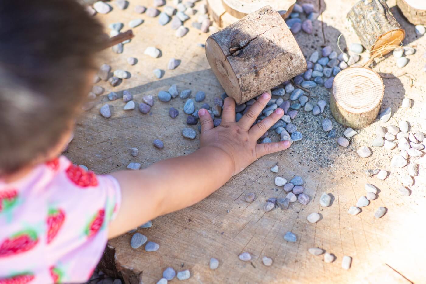 An infant’s hand feels pebbles and sand on a tree stump.