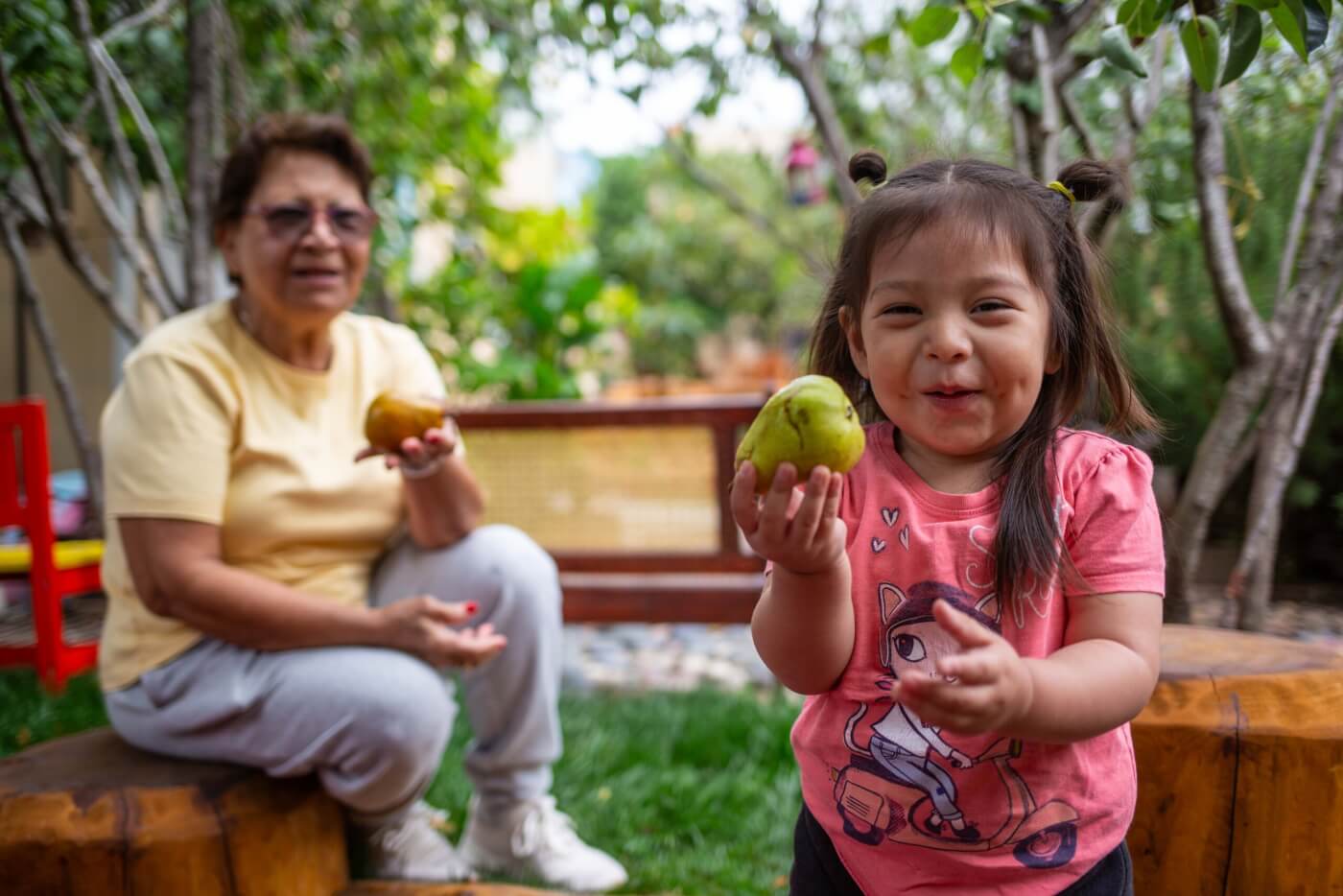 A young child holds up a fruit and smiles, while a caregiver looks on in the background.