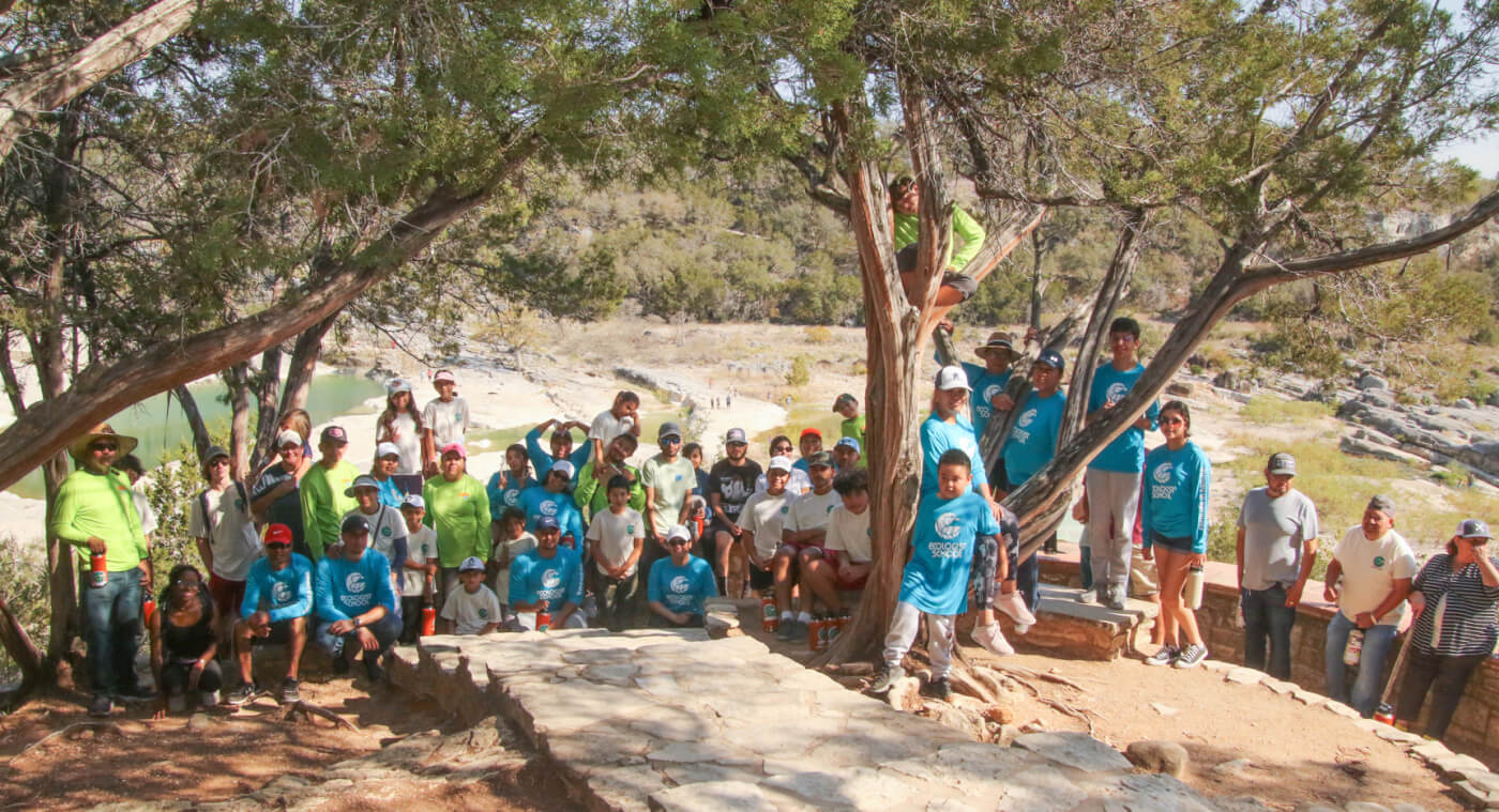 group of people with matching blue and green shirts smiling at camera with trees, water, and rocks in the background