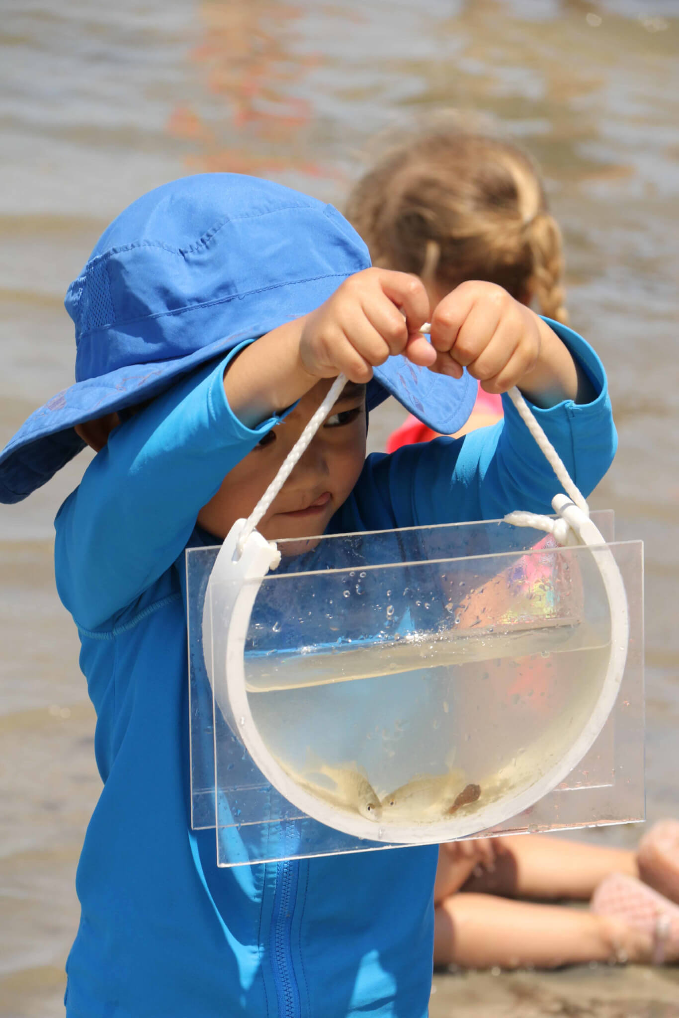 small child in blue hat and sun protection jacket holding small tank with fish and water