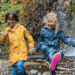 Two children hold hands and play in the rain. One child has braids and a yellow rain jacket. One child has blond hair and a blue patterned rain jacket.
