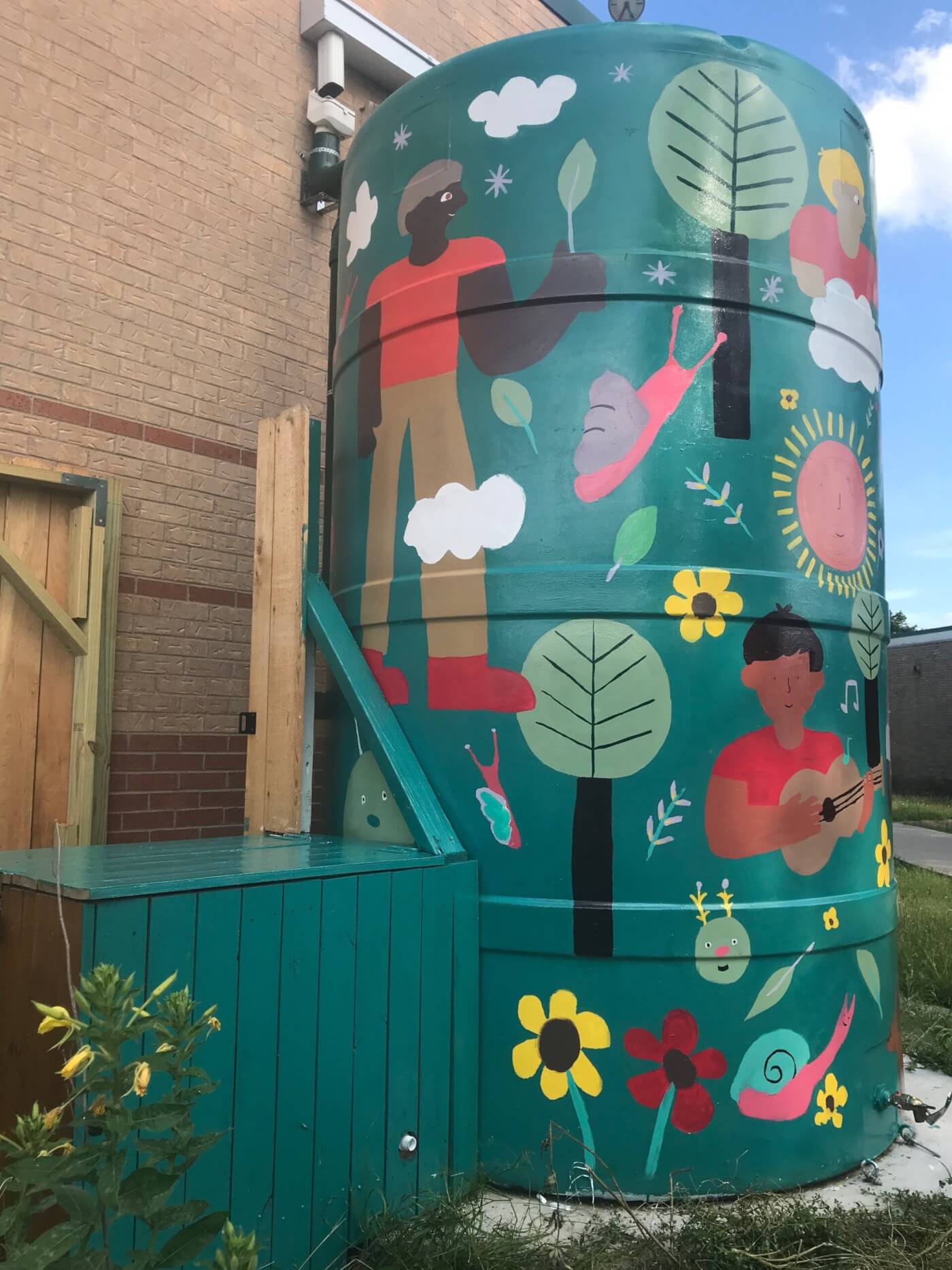 A large green rainwater cistern at Barrington Elementary School decorated with a colorful mural. The artwork depicts people interacting with nature — one holding a leaf, another playing guitar — surrounded by trees, flowers, clouds, and smiling suns and snails. The cistern sits beside a brick building with a wooden enclosure, symbolizing the school’s commitment to sustainability and outdoor learning.