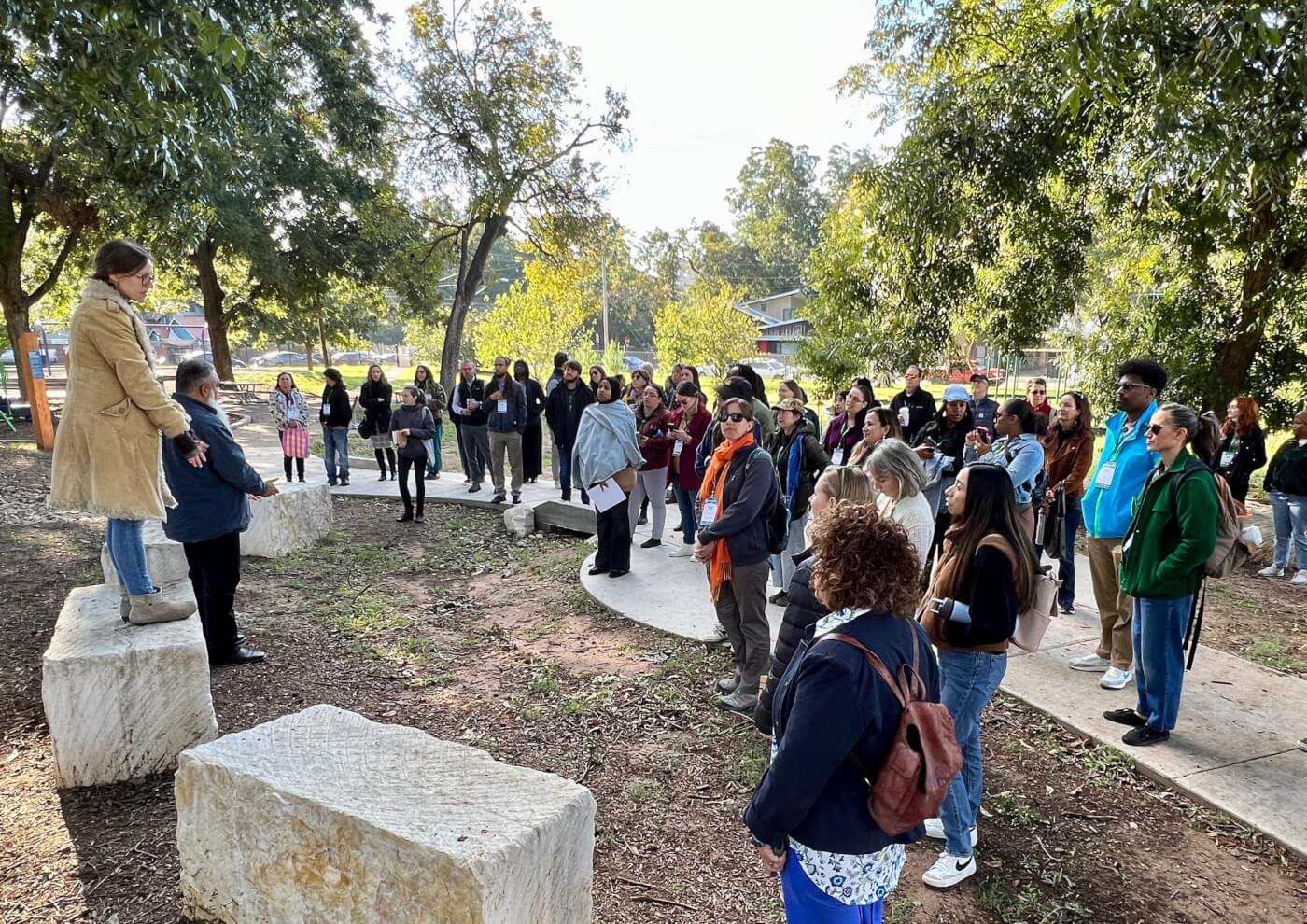 people standing on a sidewalk facing a woman who is standing on a cement block talking with the whole group.