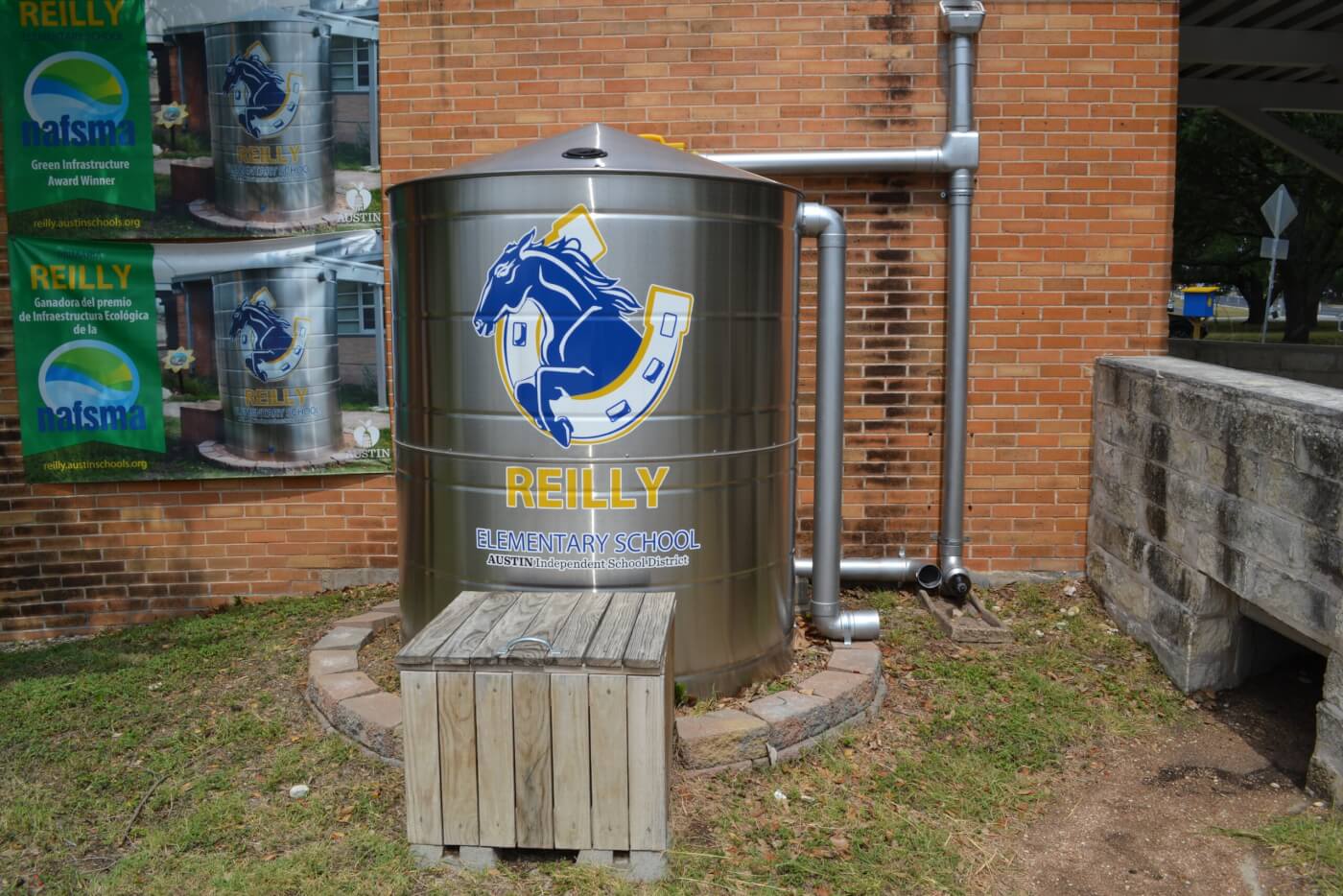 A large silver rainwater cistern with the Reilly Elementary School logo, featuring a blue horse and yellow horseshoe, installed beside a brick school building. The cistern is connected to metal downspouts for stormwater collection, with a small wooden box in front. A banner on the wall highlights the school’s NAFSMA Green Infrastructure Award for its stormwater project.