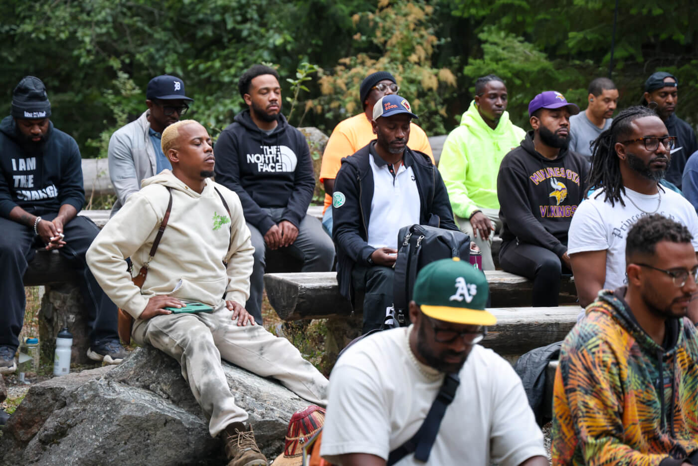 A group of 13 Black men participating in the Boyz N The Wood retreat sit on log benches and rocks. Their eyes are closed in reflection.
