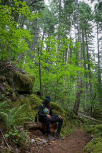A man sits quietly on a log in a mossy forest with his head bowed.