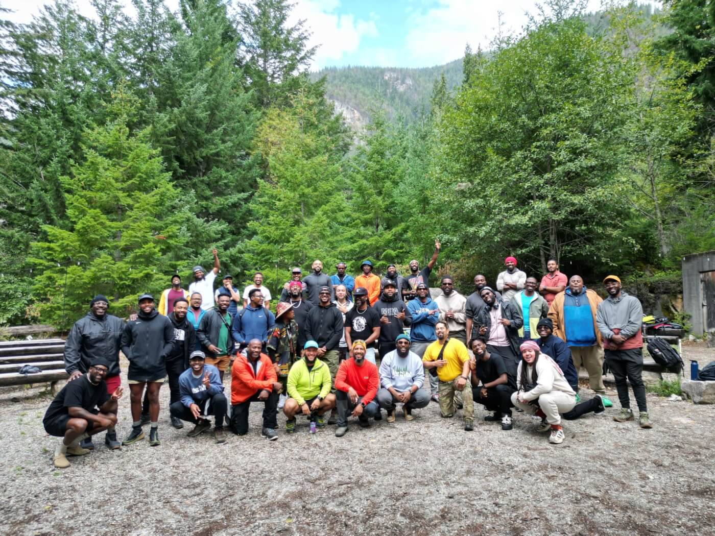 A group of Boyz N the Wood retreat participants pose in a rustic amphitheater, with forest and mountains in the background.