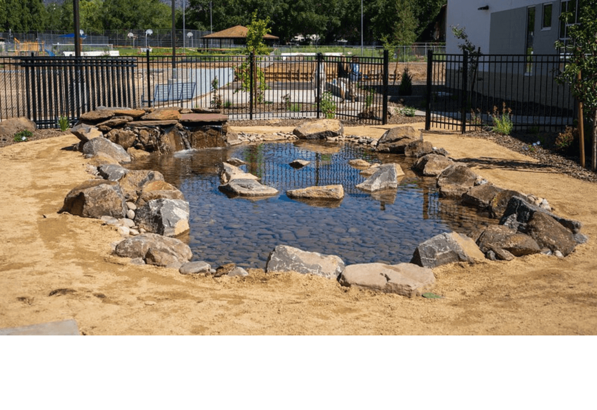 An element of Killip Elementary’s green schoolyard designed to support water retention, featuring a small rock-lined pond with a gentle waterfall and surrounding sandy terrain.