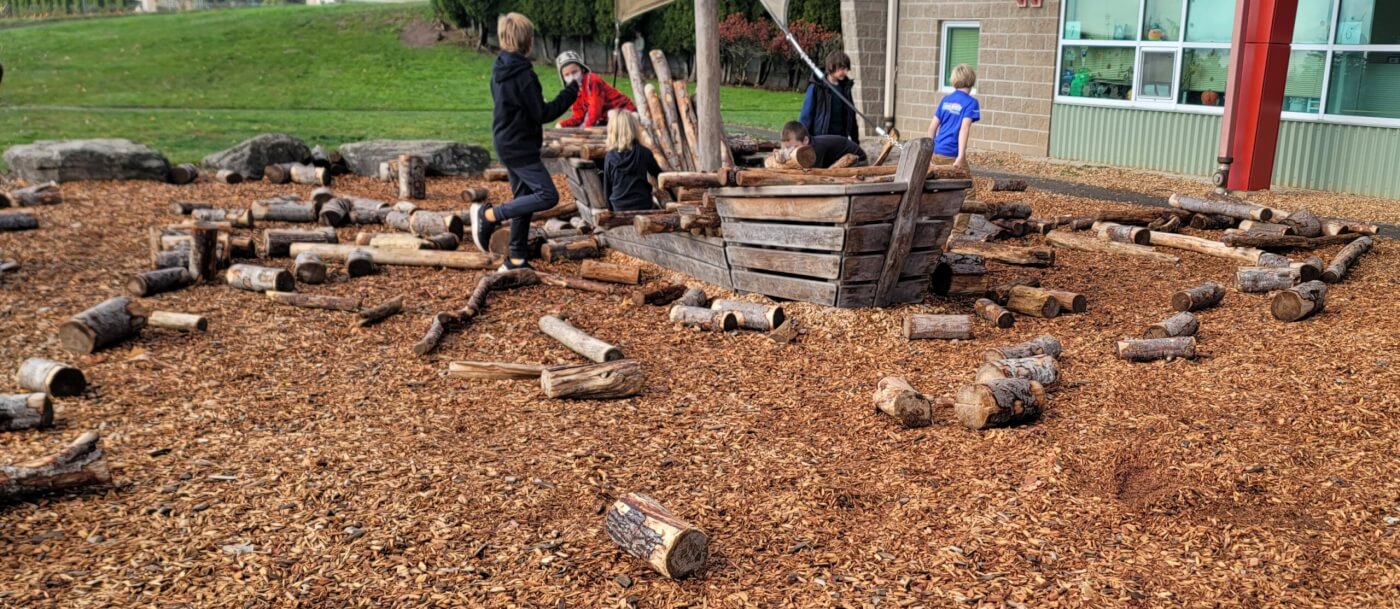 Children play and build with loose logs around a wooden boat structure in a nature-based school playground.