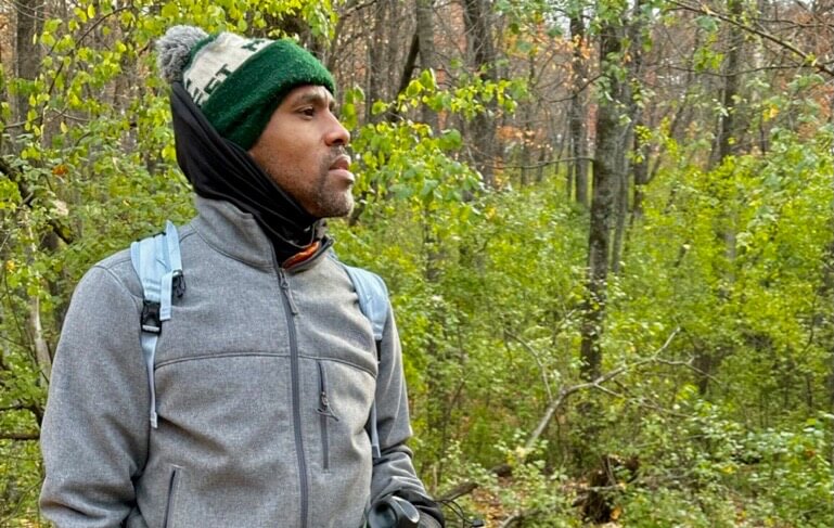 Carlos Steward, wearing a gray jacket and green beanie, looks out at the forest.