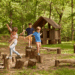 Three young children playing on wooden stumps and logs in a forest playground near a small wooden hut.