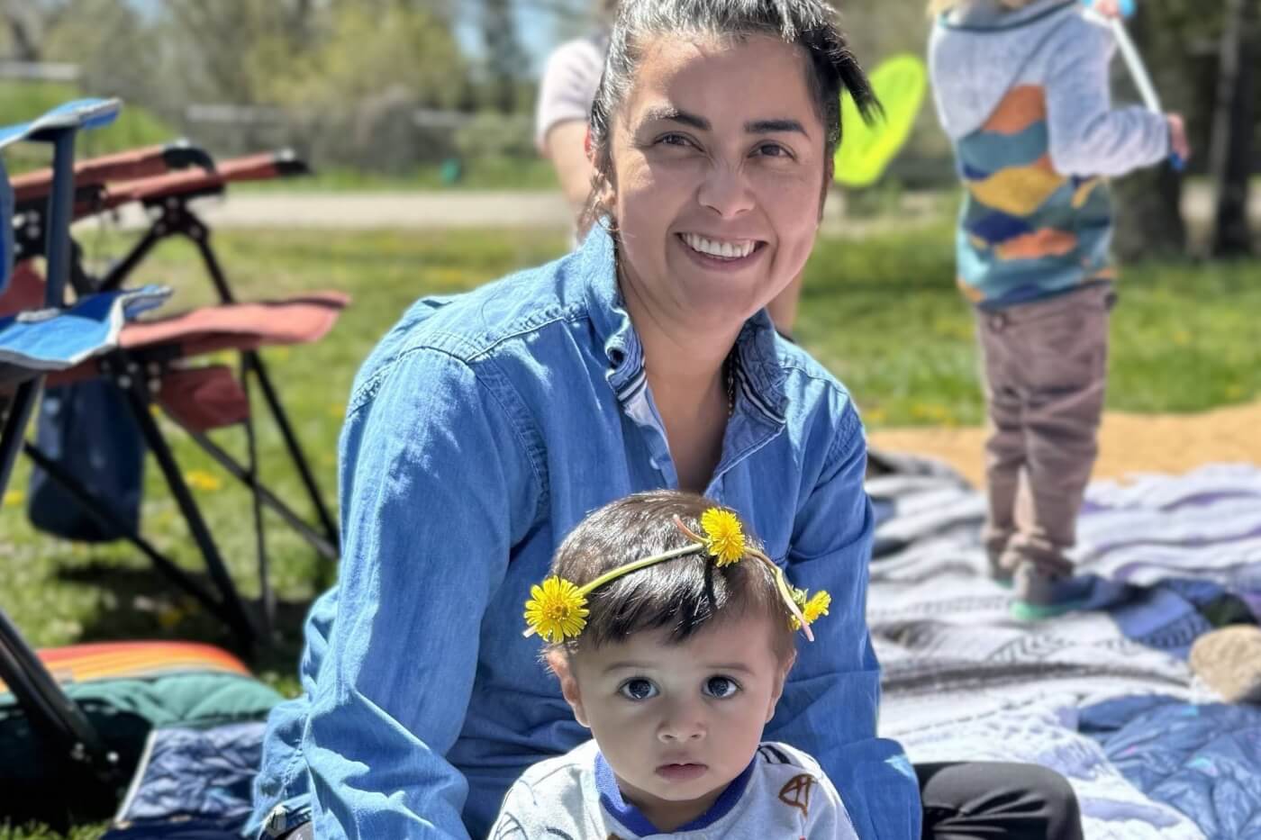 A group of workers in helmets and waders carry a large metal structure across a grassy area. A woman smiles while sitting on a blanket outdoors with a young child wearing a dandelion flower crown.