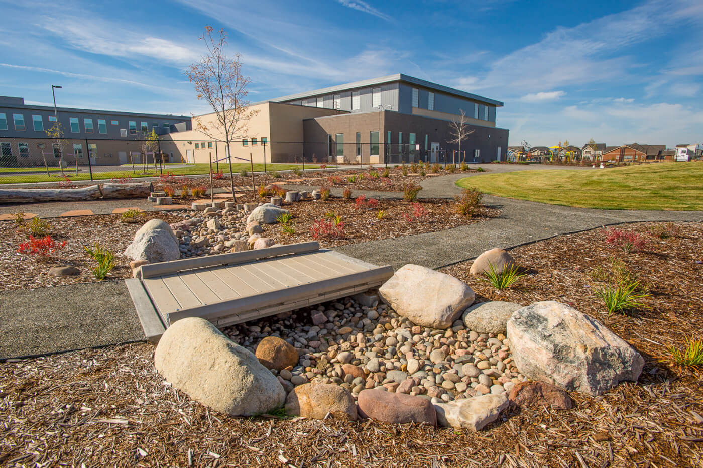 A walking bridge crosses over a shallow ditch filled with stones, ringed with larger rocks. The surrounding area is mulched and has newly planted trees and small shrubs.