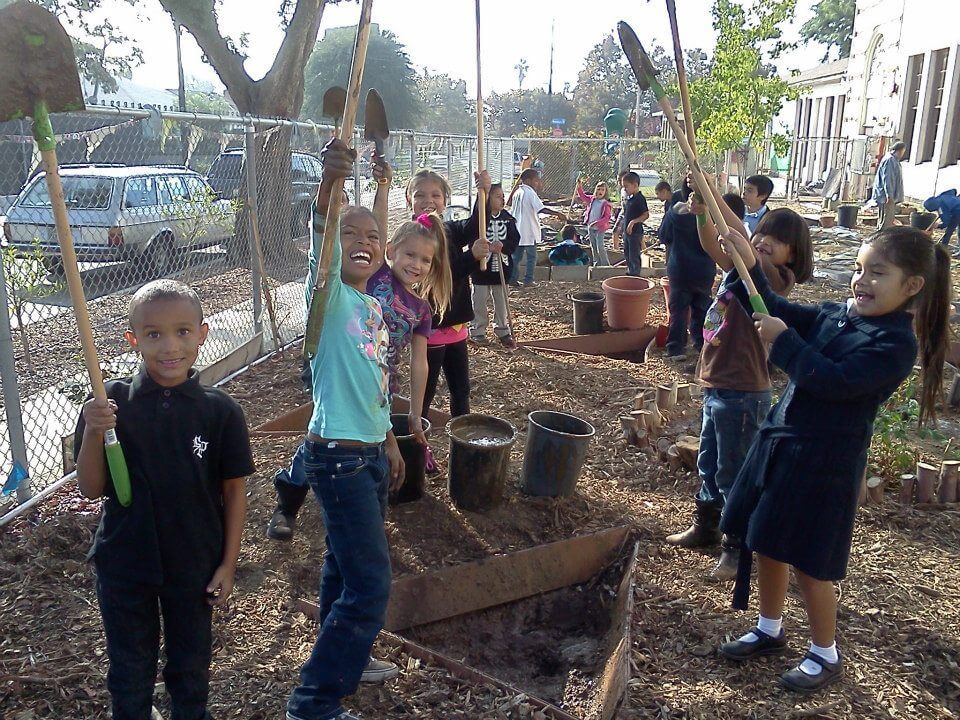 Children smiling with shovels raised in the air next to small dug out shapes for planting.