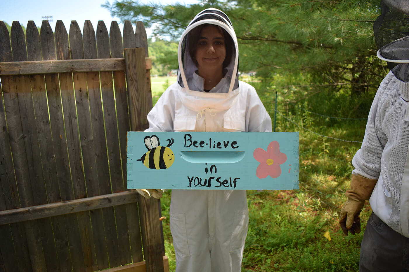 A student in a beekeeping suit carries a piece of a wooden beehive, which has been painted to read: “Bee-live in yourself.”