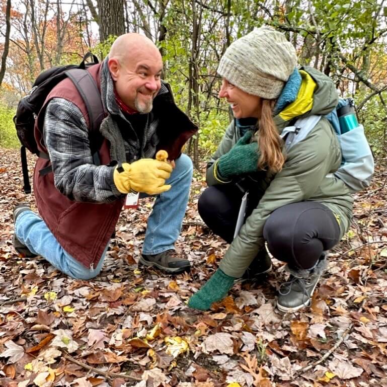 Two teachers crouch on the forest floor, covered with fallen leaves, to look at a mushroom growing along the trail.