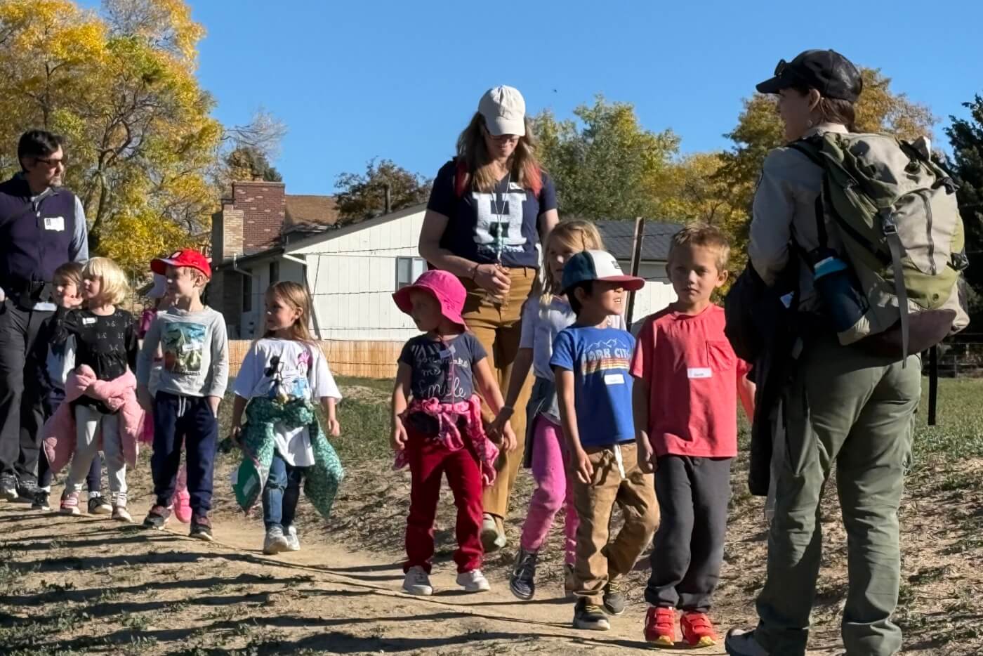 A group of young children walk along a dirt path with adults during an outdoor field trip.