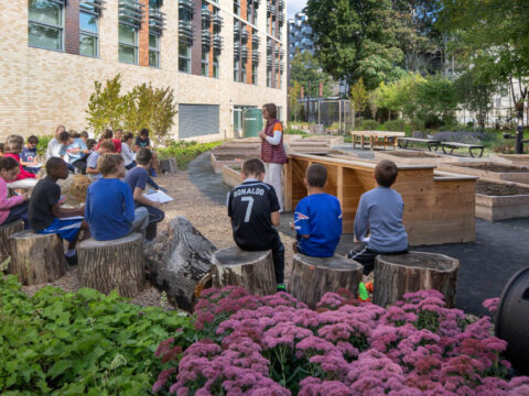 Students sit on log stools in an outdoor classroom garden beside a school building, listening to a teacher near raised planting beds surrounded by trees and flowers.