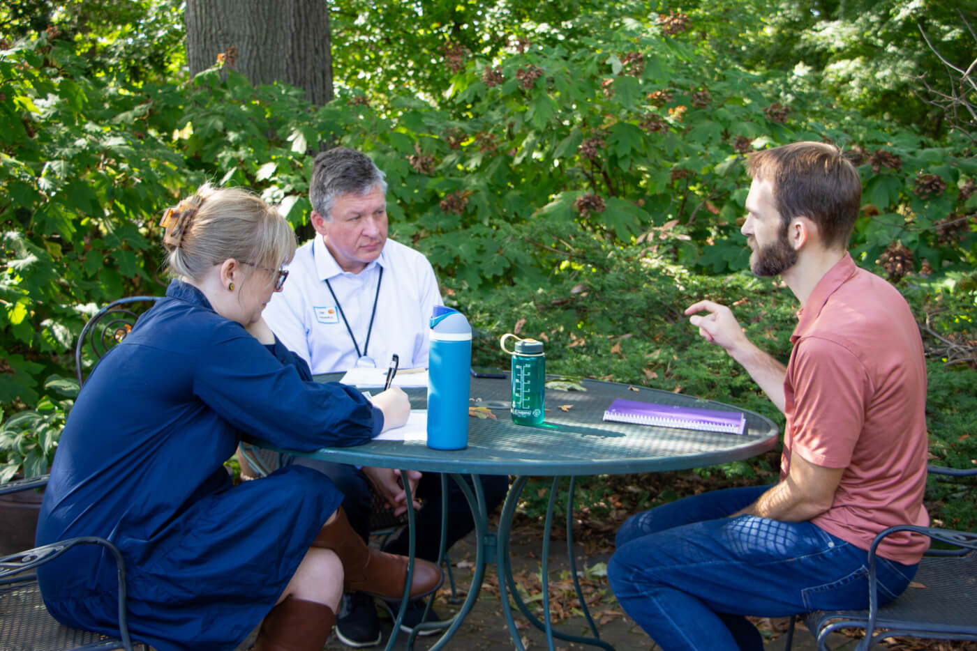 Three people sit at an outdoor table surrounded by greenery, talking while one person writes notes.