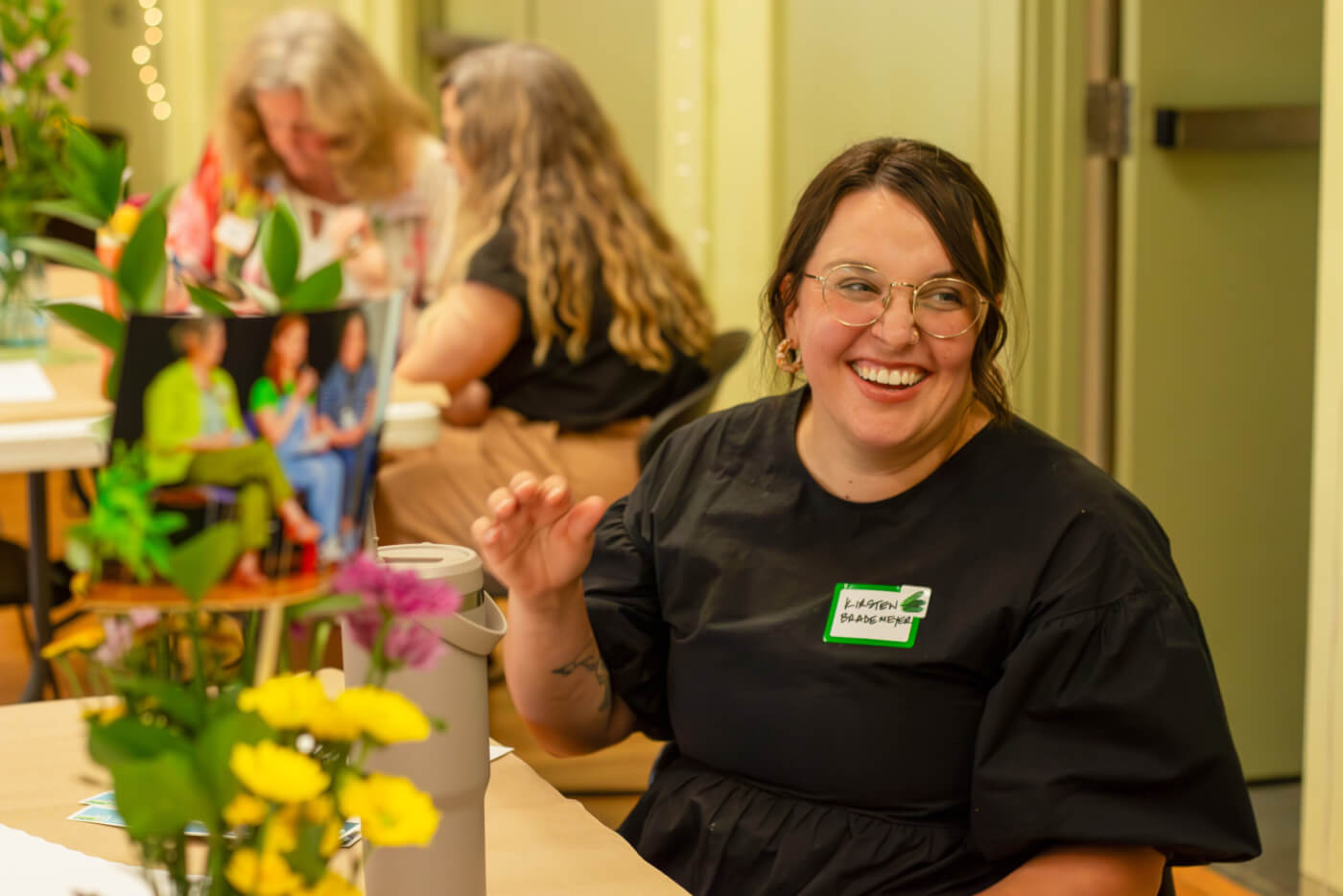A woman wearing glasses and a name tag smiles while sitting at a table with flowers, as others talk in the background.