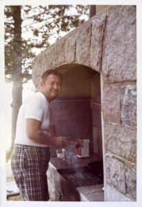 A man in plaid shorts smiles while cooking on a stone grill.