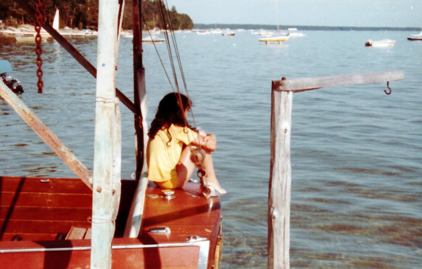 A young girl sits on the back of a wooden boat, looking out at a lake contemplatively.