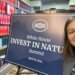 Woman taking a selfie in front of a dark blue poster board at the White House that says "White House Invest In Nature Summit"