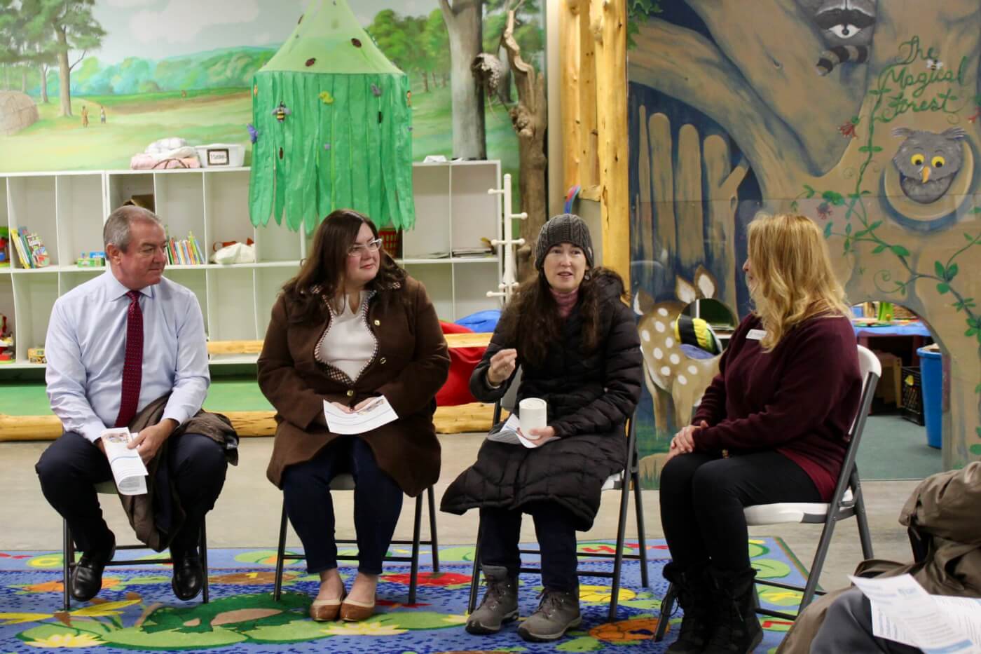 Four adults seated in a circle having a discussion inside a colorful, child-themed room.