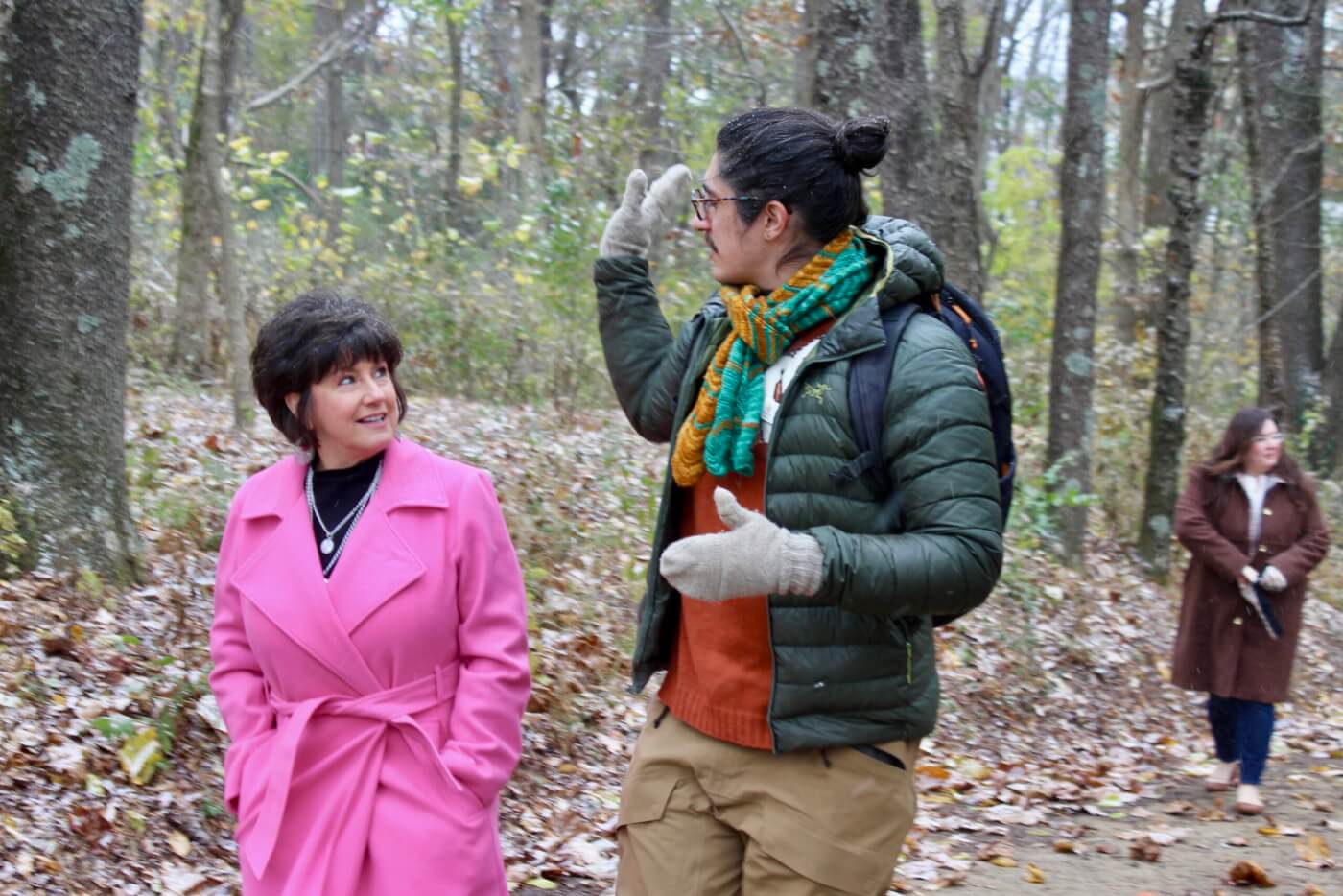 Two adults walking and talking on a forest path while another person walks behind them.
