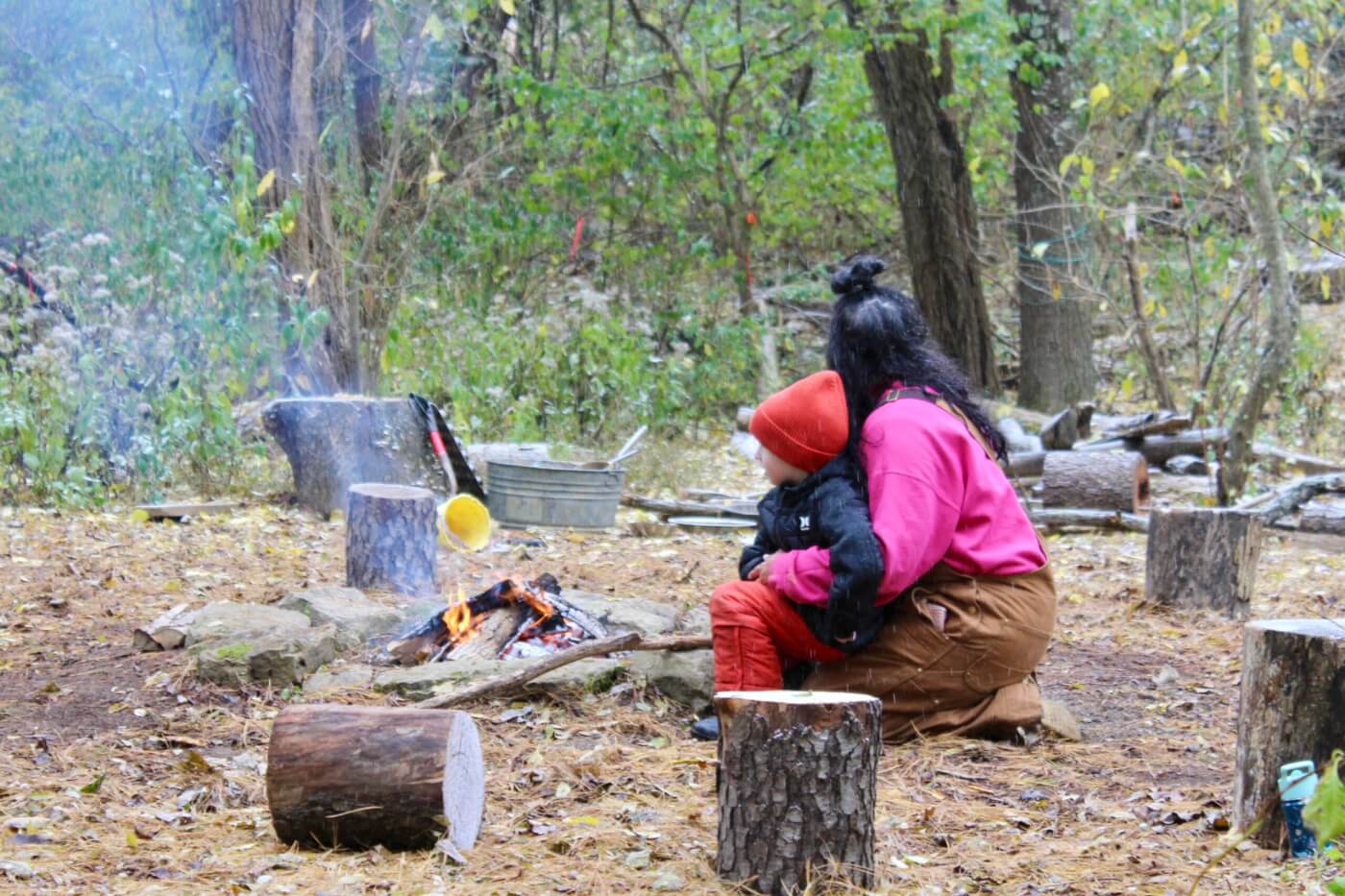 Adult and young child sitting together by a small campfire in a wooded area.
