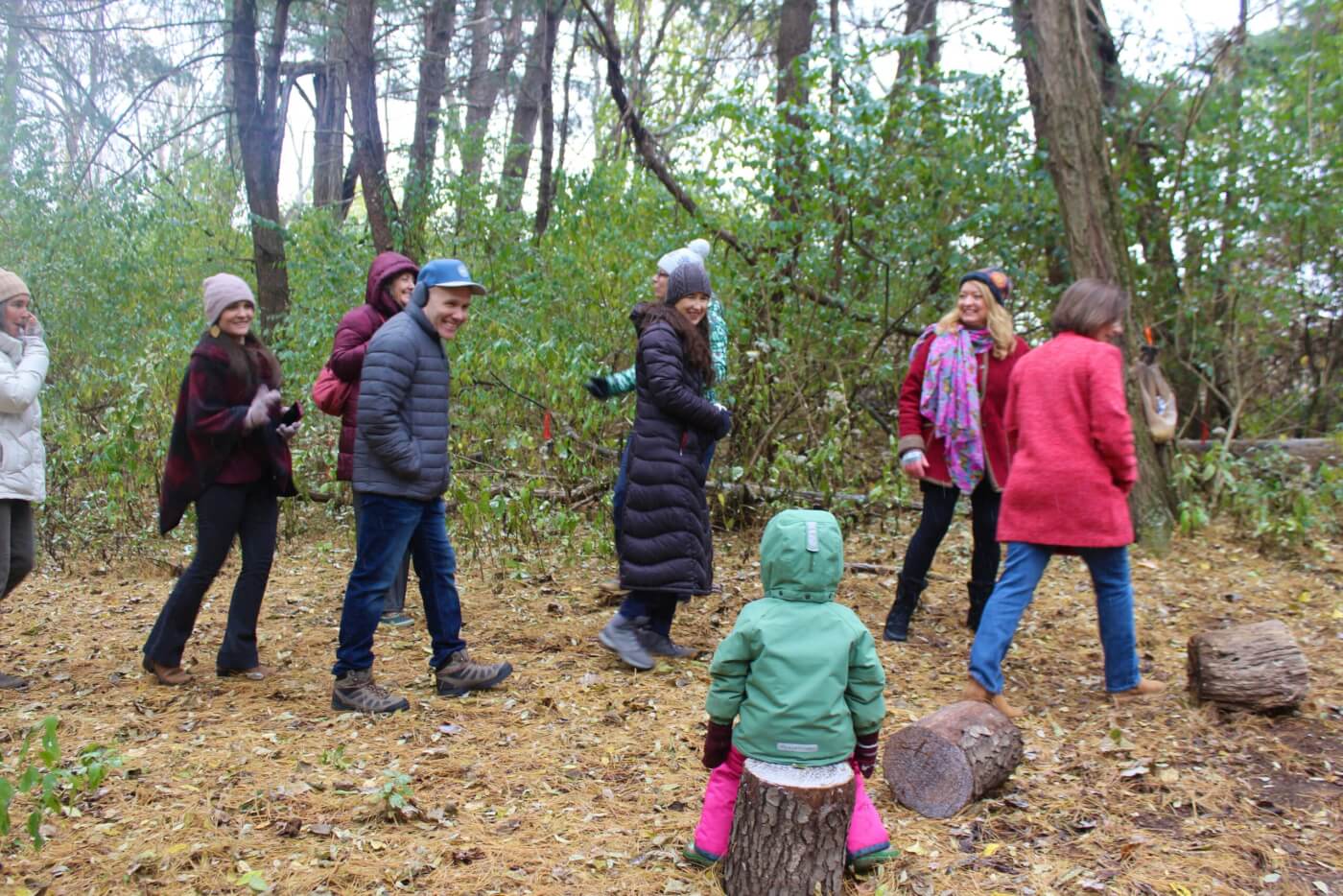 Group of adults walking together through a wooded area while a small child sits on a tree stump in the foreground.