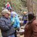 Adult holding a child while talking with another adult in a wooded area, with children playing on tree stumps behind them.
