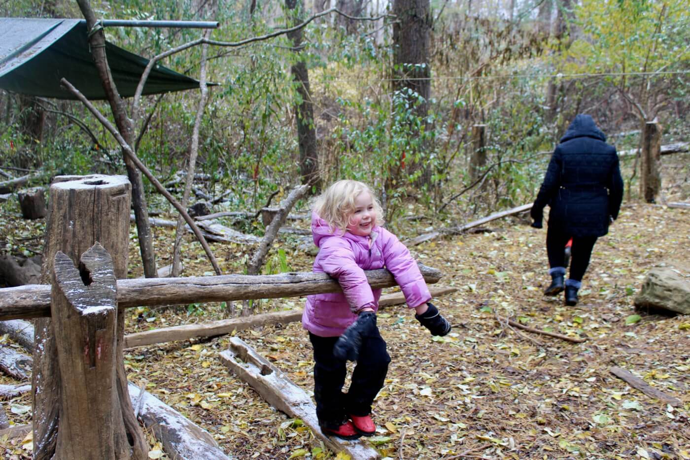 Young child in a pink coat balancing on a wooden beam in a wooded area while an adult walks away in the background.