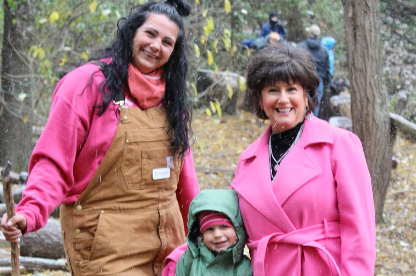 Two smiling adults and a young child standing together in a wooded area during light snowfall.