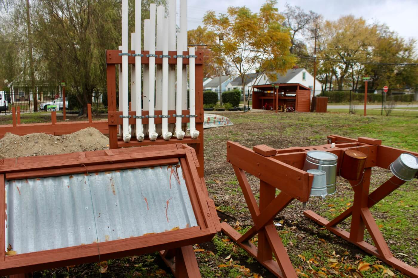 Outdoor play area with wooden sensory structures, metal panels, pipes, and buckets set in a grassy yard.