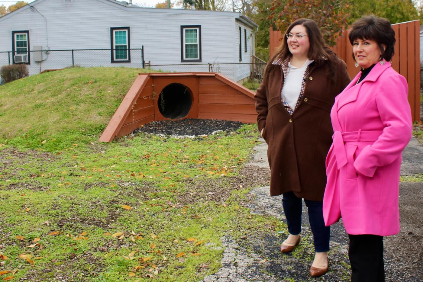 Two adults standing outdoors near a small wooden tunnel structure and grassy area.
