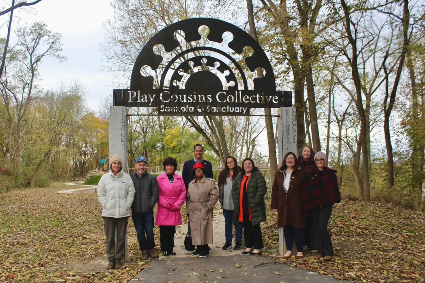 Group of adults standing together beneath a sign reading “Play Cousins Collective” at a wooded outdoor site.