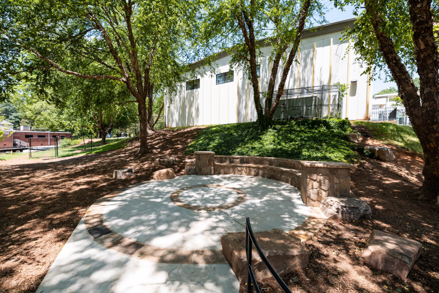 A shaded outdoor gathering space with a circular concrete pad and stone seating sits beneath mature trees beside a school building.