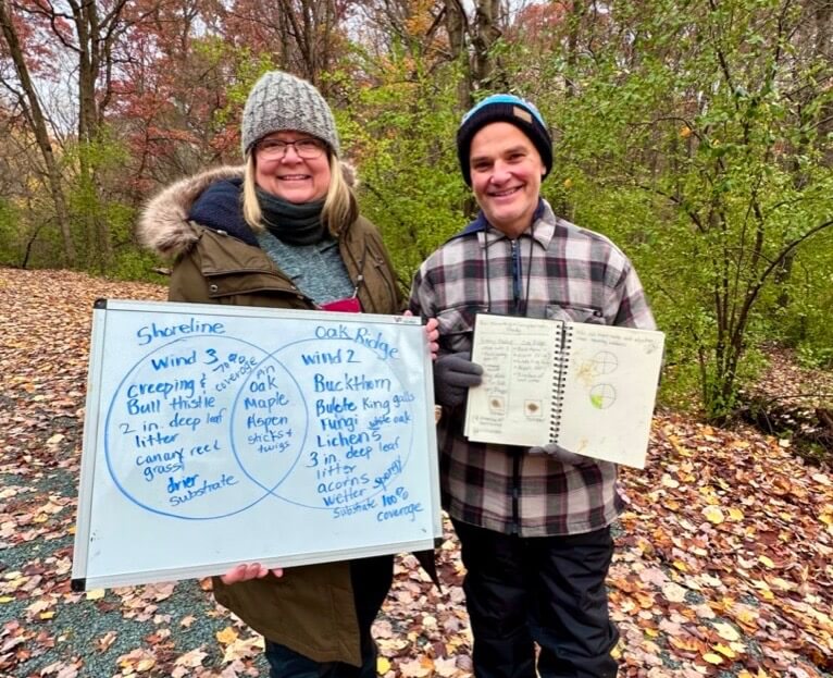 Two teachers pose for the camera. One holds a white board with a venn diagram drawn on it. The other holds a nature journal filled with notes.