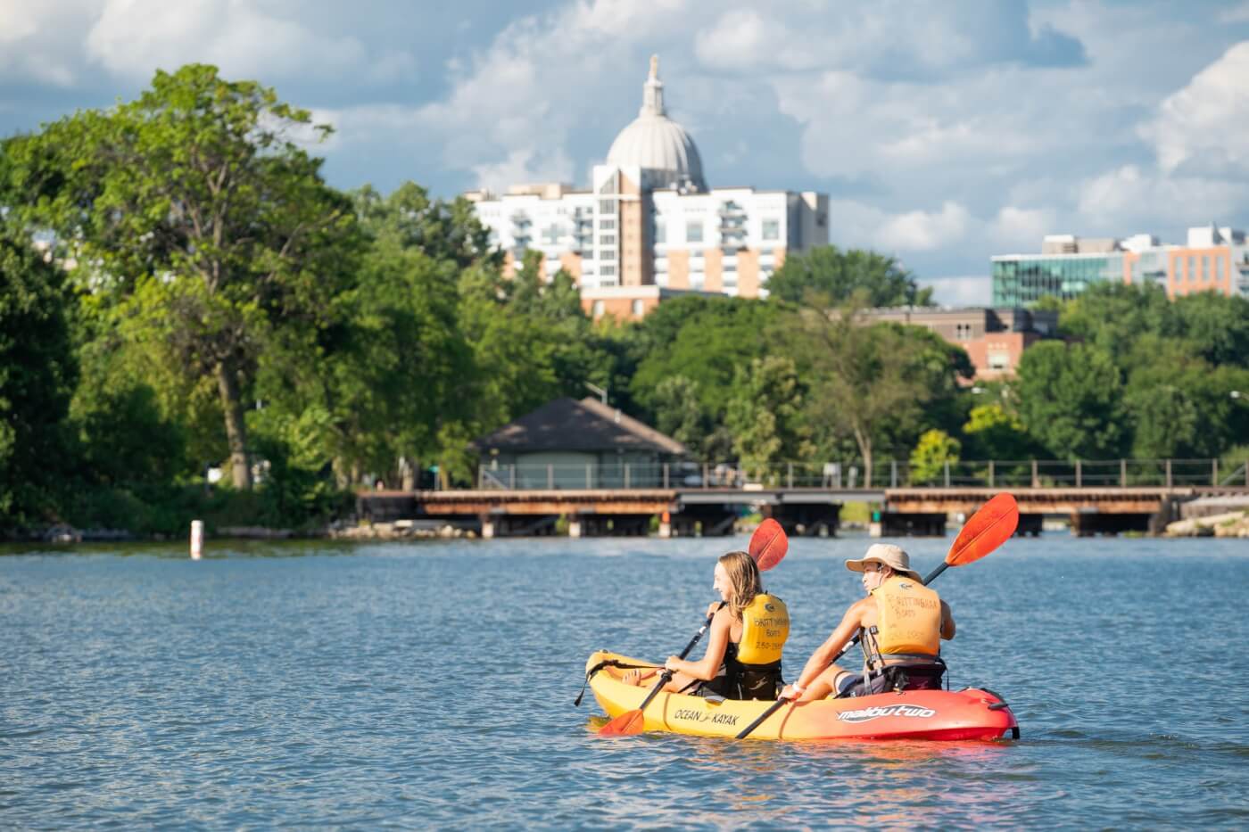 Two people in a duo kayak paddle on a lake in front of an urban setting, including the distant capital building.