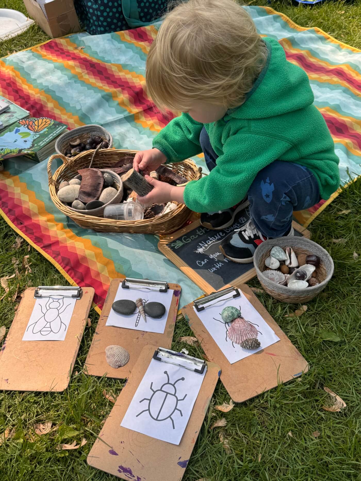 A young boy in a green sweatshirt plays with shells and rocks outside on a blanket surrounded by sheets of paper where kids can use crayons to color inside outlines of bugs
